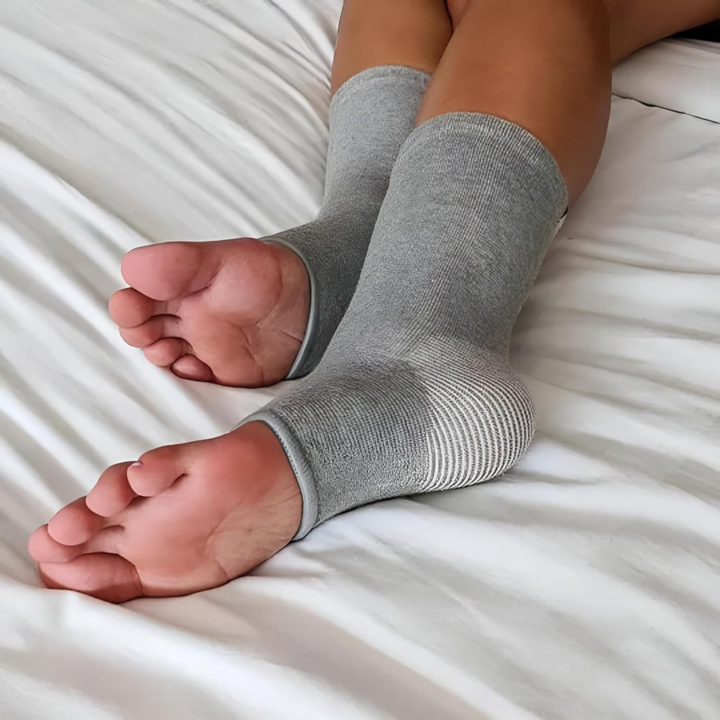 Person wearing gray ankle compression sleeves, relaxing on white bed, IndianChic wellness accessory